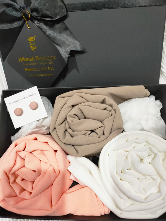 A premium gift box from Hikmah Boutique containing three neatly rolled hijabs in peach, beige, and white, along with a pair of matching hijab pins. The elegant black box is adorned with a black ribbon and gold-foil branding.